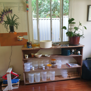 Long wooden shelf under a window inside, shelf has various containers on it, a potted plant, and some ceramics
