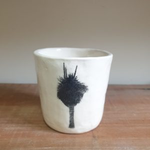 Small white porcelain cup sits on a wooden shelf. The cup is decorated with a black image of a grass tree.