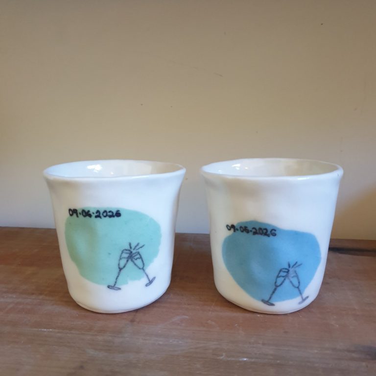 Two small white porcelain cups, each with a large patch of colour - one is light green, one is light blue. They are both decorated with a line drawing of two champagne glasses clinking and the date 09.06.2026