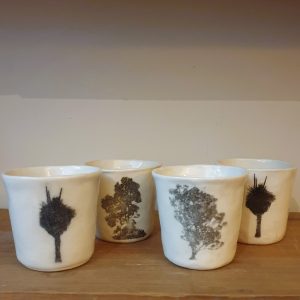 Four small white porcelain cups sit on a wooden shelf. Each is decorated with a black silhouette of a tree.