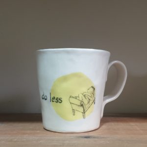 A white mug sits on a wooden shelf. The mug is decorated with a drawing of a person lying on a chaise-lounge on a yellow spotlight background and the words "do less".